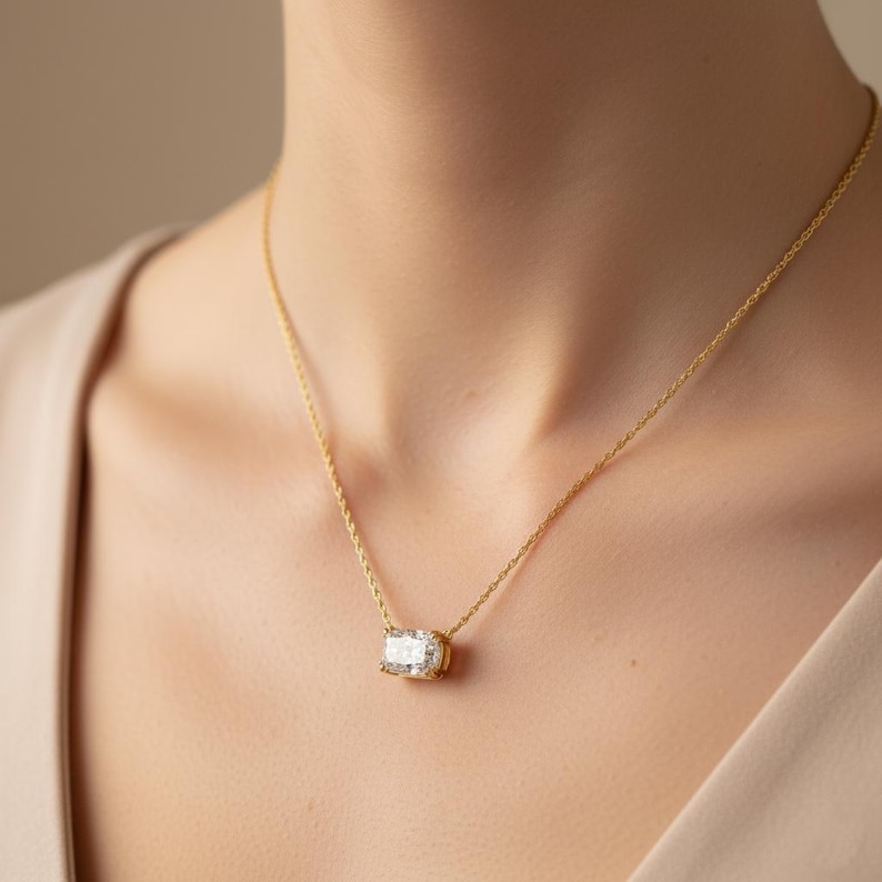 K&ouml;nnte beinhalten: Eine goldene Halskette mit einem rechteckigen, klaren Edelsteinanh&auml;nger. Der Anh&auml;nger ist in einer goldenen L&uuml;nette gefasst und h&auml;ngt an einer zarten Goldkette. Die Halskette wird am Hals einer Person getragen.