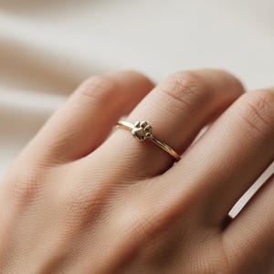 Puede incluir: Un delicado anillo de oro con un diseño de huella de pata. El anillo tiene una banda delgada y un pequeño y detallado dije de huella de pata. El anillo se muestra en un dedo, destacando su estilo elegante y minimalista. Un regalo perfecto para los amantes de las mascotas.