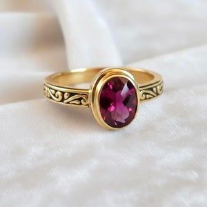 May include: A gold ring with an oval, deep red gemstone. The ring band features an ornate, black filigree design. The gemstone is set in a gold bezel, and the ring is displayed on a white surface.