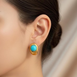 Könnte beinhalten: Goldfarbene Ohrstecker mit einem leuchtenden türkisfarbenen Stein. Die Ohrringe haben eine abgerundete, ovale Form, und der türkisfarbene Stein ist in einen polierten Goldrahmen gefasst. Das Design ist schlicht und elegant, für den Alltag geeignet.