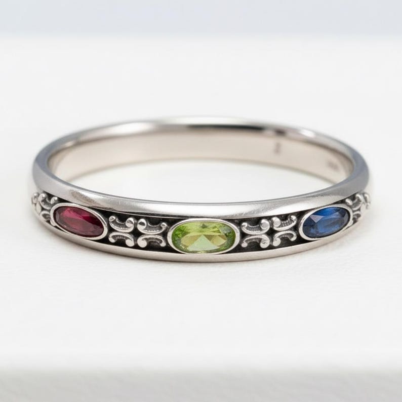 Puede incluir: Un anillo de plata con tres piedras preciosas ovaladas: roja, verde y azul. El anillo presenta un dise&ntilde;o ornamentado entre las piedras preciosas. Las piedras preciosas est&aacute;n engastadas en una banda de plata. Es una joya.