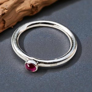 Puede incluir: Un anillo de plata con una pequeña gema redonda de color rojo intenso. El anillo tiene un diseño sencillo, con una banda lisa y pulida. La gema está engastada en un bisel. El anillo se muestra sobre una superficie gris.