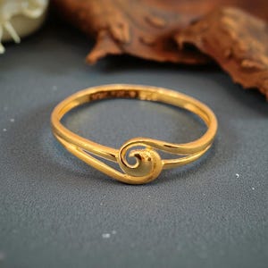 May include: A gold ring with a distinctive swirl design. The ring has a double band that curves into a spiral at the front. The ring is set against a dark grey background.