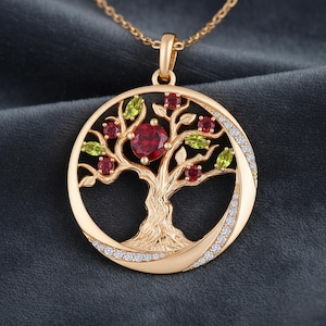 Könnte beinhalten: Goldfarbene Anhänger-Halskette mit einem Lebensbaum-Design. Der runde Anhänger zeigt einen Baum mit roten und grünen Edelsteinen. Eine halbmondförmige Form ist mit kleinen, klaren Steinen besetzt.