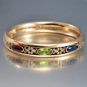 Peut inclure: Une bague dorée ornée de trois pierres précieuses ovales : rouge, verte et bleue. La bague présente un motif ornemental autour des pierres et un anneau lisse et poli. La bague est probablement en or.