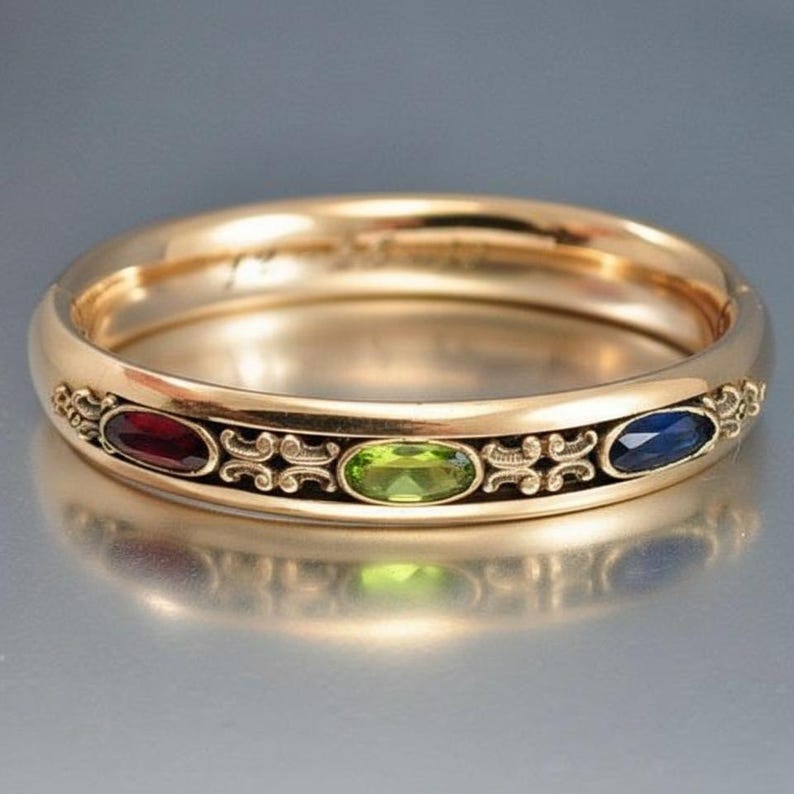 Puede incluir: Un anillo dorado con tres piedras preciosas ovaladas: roja, verde y azul. El anillo tiene un dise&ntilde;o ornamental alrededor de las piedras y una banda lisa y pulida. El anillo es probablemente de oro.