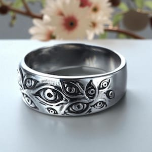 May include: A silver ring with an eye pattern. The ring has a series of embossed eyes around the band. The ring is made of a shiny, metallic material and has a smooth, polished finish. The ring is a wide band style.