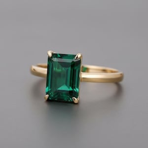 以下が含まれることがあります： 長方形のエメラルドカットの緑色の宝石をあしらったゴールドリング。リングは、滑らかで光沢のあるゴールドバンドを備えたシンプルなデザインです。宝石は鮮やかで深い緑色です。婚約指輪。