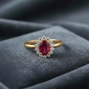 Könnte beinhalten: Ein goldener Ring mit einem ovalen Rubin-Mittelstein, umgeben von einem Kranz aus kleinen, runden Diamanten. Der Ring sitzt auf einem schlichten, polierten Goldband und wird auf einem grauen Samtstoff präsentiert.