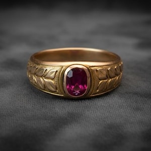 May include: A gold-colored ring with a large, oval-shaped, deep pink gemstone at the center. The band features a leaf design on both sides of the gemstone. The ring is set against a dark gray background.