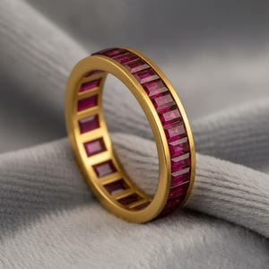 May include: A gold ring with a band of rectangular, deep red gemstones. The ring's gold band is smooth and polished, and the gemstones are set closely together, creating a continuous line of color. The ring is displayed on a soft, gray surface.