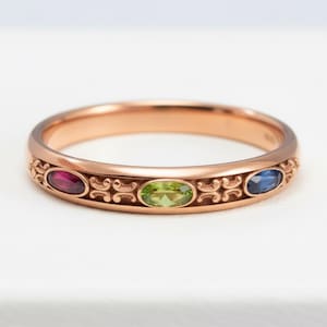 Puede incluir: Anillo de oro rosa con tres piedras preciosas ovaladas: un rub&iacute;, un peridoto y un zafiro. El anillo presenta un dise&ntilde;o ornamental de volutas repetidas a lo largo de la banda. Las piedras preciosas est&aacute;n dispuestas en fila.