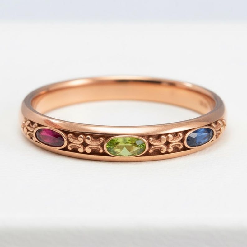 Puede incluir: Anillo de oro rosa con tres piedras preciosas ovaladas: un rub&iacute;, un peridoto y un zafiro. El anillo presenta un dise&ntilde;o ornamental de volutas repetidas a lo largo de la banda. Las piedras preciosas est&aacute;n dispuestas en fila.