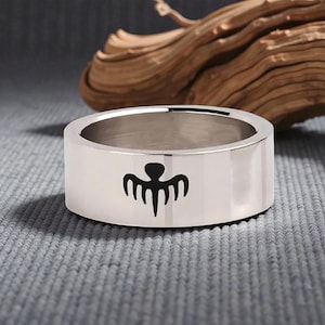 Peut inclure: Bague argentée avec une bande plate et un motif graphique noir. L'anneau est posé sur un fond gris avec du bois en arrière-plan. Le design de l'anneau est une forme symétrique stylisée.