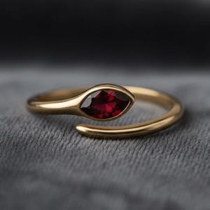 May include: A gold-colored ring with a marquise-cut, deep red gemstone. The ring's band curves around to meet the gemstone setting, creating a unique, open design. The ring is set against a gray, textured background.