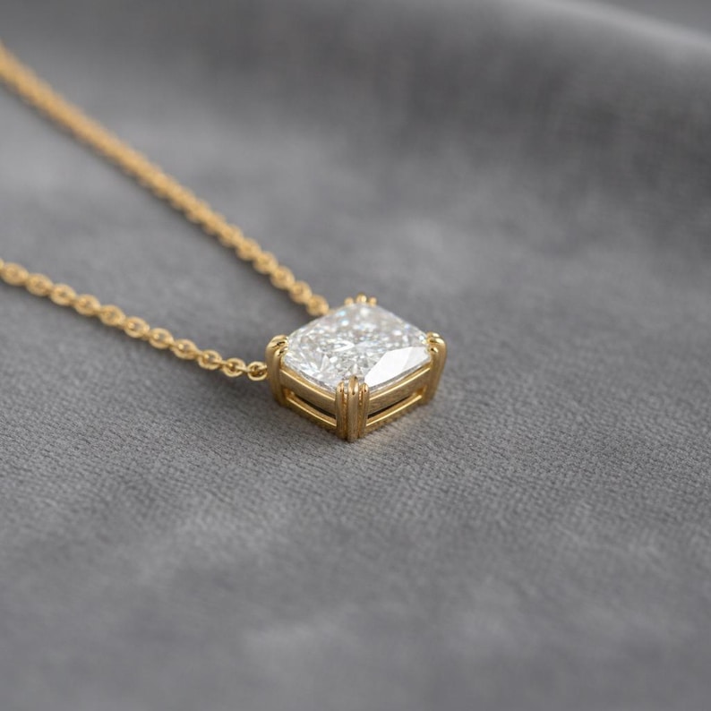 K&ouml;nnte beinhalten: Eine goldene Halskette mit einem gro&szlig;en, quadratisch geschliffenen Diamantanh&auml;nger. Der Diamant ist in einer goldenen L&uuml;nette gefasst und h&auml;ngt an einer zarten Goldkette. Die Halskette wird auf einer grauen Oberfl&auml;che pr&auml;sentiert.