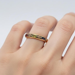 Puede incluir: Un anillo de oro con cuatro piedras preciosas ovaladas en rojo, verde y azul. El anillo tiene un borde decorativo de esmalte negro con detalles florales. El anillo est&aacute; en un dedo.