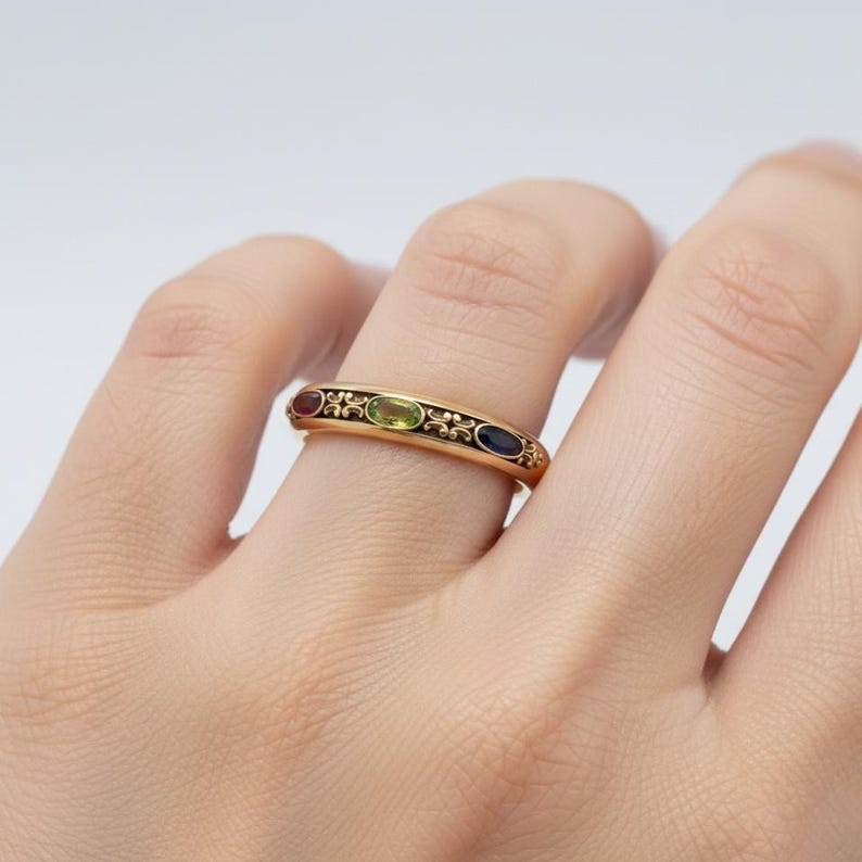 Puede incluir: Un anillo de oro con cuatro piedras preciosas ovaladas en rojo, verde y azul. El anillo tiene un borde decorativo de esmalte negro con detalles florales. El anillo est&aacute; en un dedo.