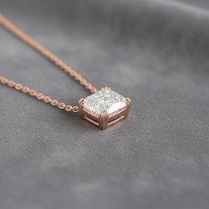 K&ouml;nnte beinhalten: Eine ros&eacute;goldene Halskette mit einem quadratisch geschliffenen Diamantanh&auml;nger. Der Diamant ist in einer Vier-Krappen-Fassung gefasst und h&auml;ngt an einer zarten Kette. Die Halskette wird auf einer grauen Oberfl&auml;che pr&auml;sentiert.
