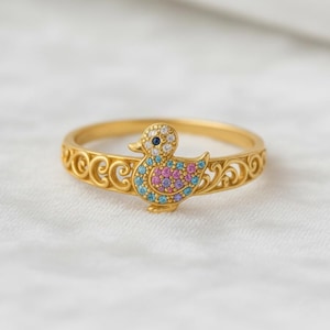 Puede incluir: Un anillo dorado con un diseño de pato. El pato está adornado con gemas azules, rosas y blancas. La banda del anillo tiene un patrón ornamental y ondulado. El ojo del pato es una pequeña gema azul oscuro.