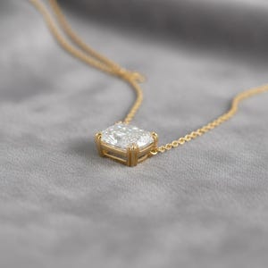 K&ouml;nnte beinhalten: Eine goldene Halskette mit einem quadratisch geschliffenen, klaren Edelsteinanh&auml;nger. Der Anh&auml;nger ist in einer goldenen L&uuml;nettenfassung gefasst und h&auml;ngt an einer zarten Goldkette. Der Hintergrund ist ein weicher, grauer Stoff.