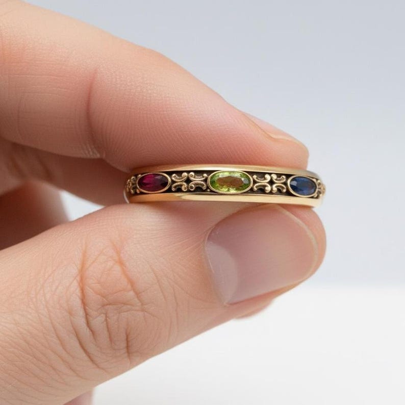 Puede incluir: Un anillo de oro con tres piedras preciosas ovaladas en rojo, verde y azul. El anillo tiene un dise&ntilde;o decorativo de filigrana negra entre las piedras. El anillo se sostiene entre los dedos.
