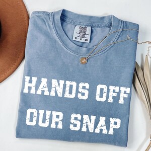 May include: A blue Comfort Colors t-shirt with the text "HANDS OFF OUR SNAP" in white block letters. A gold necklace with a pendant is draped across the shirt. A brown hat is partially visible in the upper left corner.