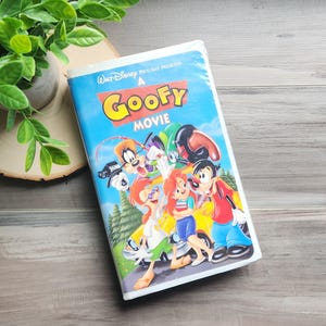 Peut inclure: Une cassette VHS du film "A Goofy Movie" de Disney, avec une image de dessin animé colorée de Dingo et d'autres personnages. La cassette est sur une surface en bois avec une plante verte en arrière-plan.