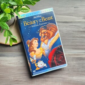 May include: A VHS tape of Disney's "Beauty and the Beast - Special Edition." The cover shows Belle and the Beast, set against a blue backdrop, with the film's title in gold. The tape rests on a wooden surface, next to some greenery.