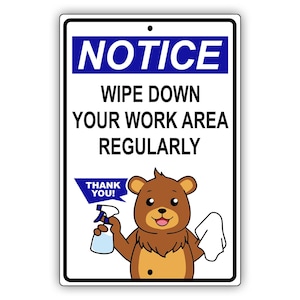 May include: A rectangular sign with a white background and black border. The sign reads "NOTICE" in blue, followed by "WIPE DOWN YOUR WORK AREA REGULARLY." A cartoon bear holds a spray bottle and a cloth. The sign also says "THANK YOU!"