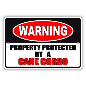 May include: Rectangular sign with a black border and a white background. The top section has a red oval with the word "WARNING" in white. The bottom section reads "PROPERTY PROTECTED BY A CANE CORSO" in black and red.