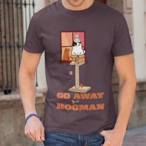 May include: A dark purple t-shirt featuring a cat-themed graphic. The graphic includes a cat tree, a window with cat silhouettes, and the text "GO AWAY HOOMAN" in orange. The shirt is a short-sleeved crew neck.