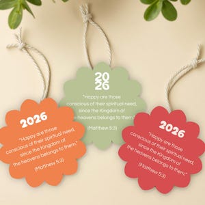May include: Three flower-shaped ornaments in orange, green, and red, each with the year "2026" in white. The ornaments have a quote from Matthew 5:3. Each ornament is suspended by a white string.