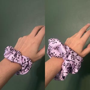 Puede incluir: Dos coleteros de satén lavanda con un estampado floral negro. Los coleteros se muestran en una muñeca sobre un fondo verde. Los coleteros están hechos de un material suave y brillante.