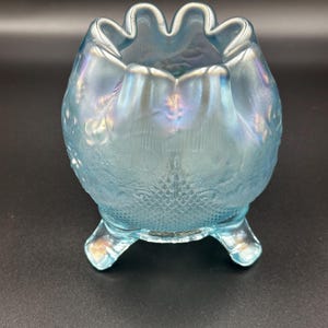 May include: A light blue, iridescent glass bowl with a ruffled rim and three small feet. The bowl has a textured, patterned surface and is likely vintage. The bowl is sitting on a black surface.
