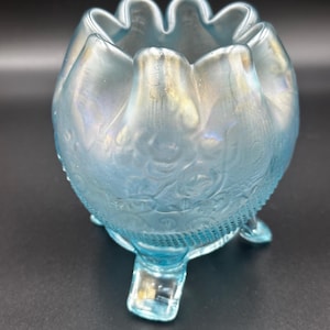 May include: A light blue, iridescent glass bowl with a ruffled rim and three small feet. The bowl has a floral pattern embossed on its surface and a textured band near the base. The bowl is approximately 4 inches in diameter.