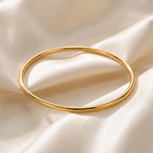 Puede incluir: Una pulsera tipo brazalete circular de oro con un acabado liso y pulido. La pulsera presenta un diseño de banda fina y uniforme que crea un aro limpio y continuo, reflejando la luz sobre su superficie metálica brillante.
