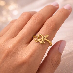 Könnte beinhalten: Ein goldfarbener Ring mit dem Buchstaben "A" in einem stilisierten Schriftzug. Der Ring wird an einem Finger getragen und zeigt sein zartes und elegantes Aussehen. Das Ringband ist dünn, und der Buchstabe "A" ist der Blickfang.