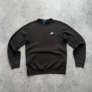May include: Black crew neck sweatshirt with the Nike logo embroidered in white on the chest. The sweatshirt has a small tag on the back of the neck. The sleeves and bottom hem have a ribbed design.