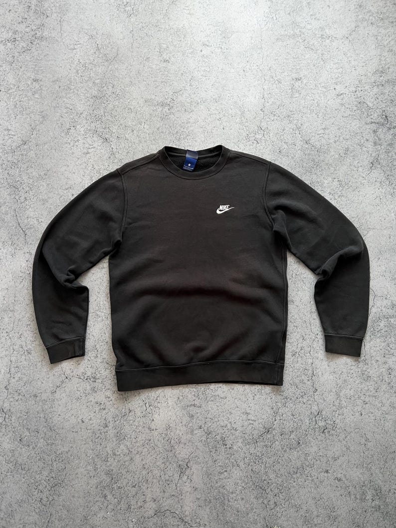 May include: Black crew neck sweatshirt with the Nike logo embroidered in white on the chest. The sweatshirt has a small tag on the back of the neck. The sleeves and bottom hem have a ribbed design.