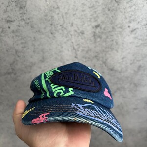 May include: A blue denim baseball cap featuring a dark blue oval patch with the text Van Dutch embroidered in black. The cap is decorated with colorful graffiti-style prints in neon green, pink, and yellow, including the text Original.