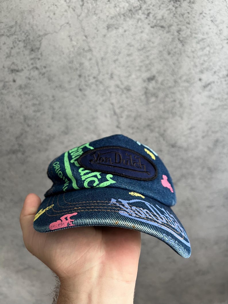 May include: A blue denim baseball cap featuring a dark blue oval patch with the text Van Dutch embroidered in black. The cap is decorated with colorful graffiti-style prints in neon green, pink, and yellow, including the text Original.