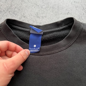 May include: Black crew neck sweatshirt with a white Nike logo on the lower right. A blue tag with white text is held up near the collar. The tag reads "S" and "MADE IN PAKISTAN".