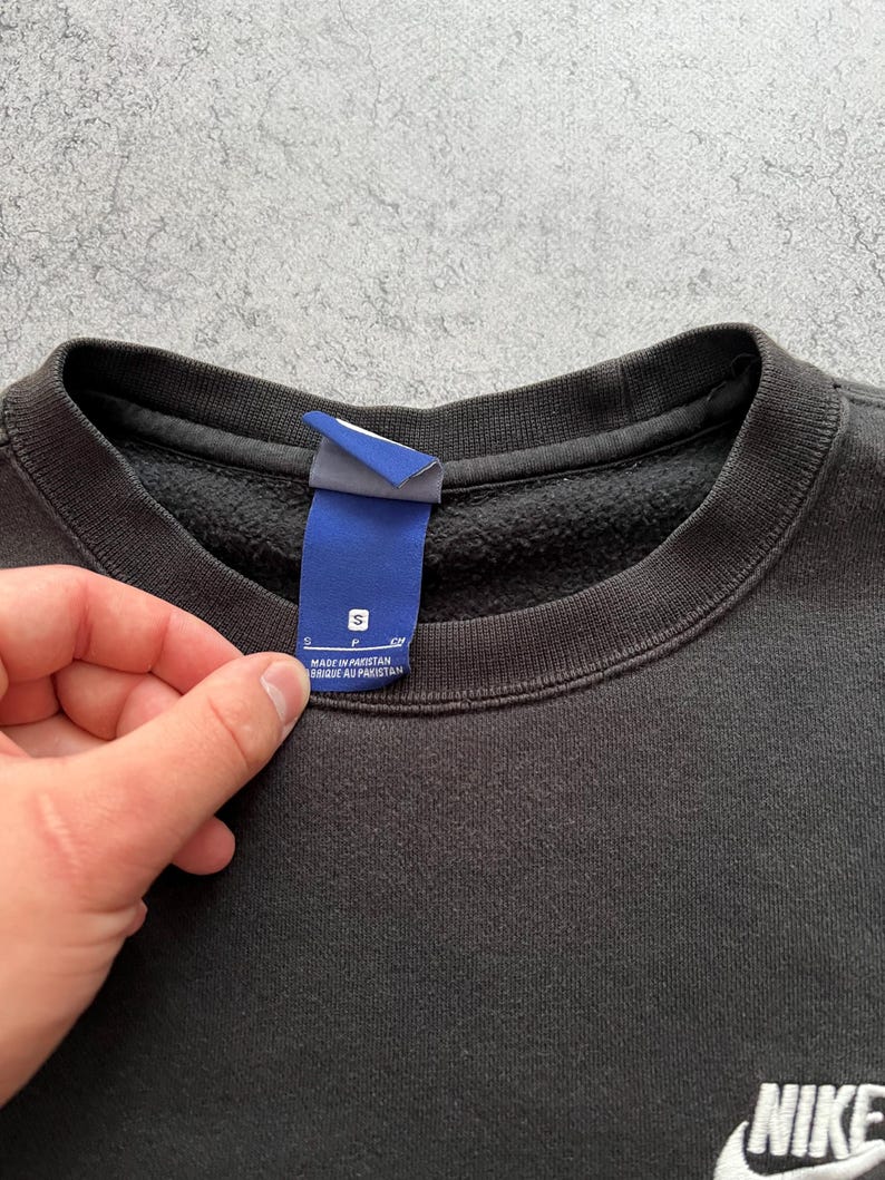 May include: Black crew neck sweatshirt with a white Nike logo on the lower right. A blue tag with white text is held up near the collar. The tag reads "S" and "MADE IN PAKISTAN".
