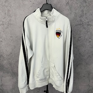 Könnte beinhalten: Weiße Jacke mit Reißverschluss, schwarzen Streifen an den Ärmeln und hohem Kragen. Mit einem deutschen Flaggenemblem und dem Text "GERMANY FIFA WORLD CUP". Mit einer Vordertasche und einem Reißverschluss.