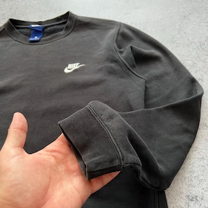 May include: Dark gray crew neck sweatshirt with the Nike logo embroidered in white on the chest. The sweatshirt has long sleeves and a ribbed cuff. The fabric appears to be slightly faded, giving it a vintage look. The collar has a blue tag.