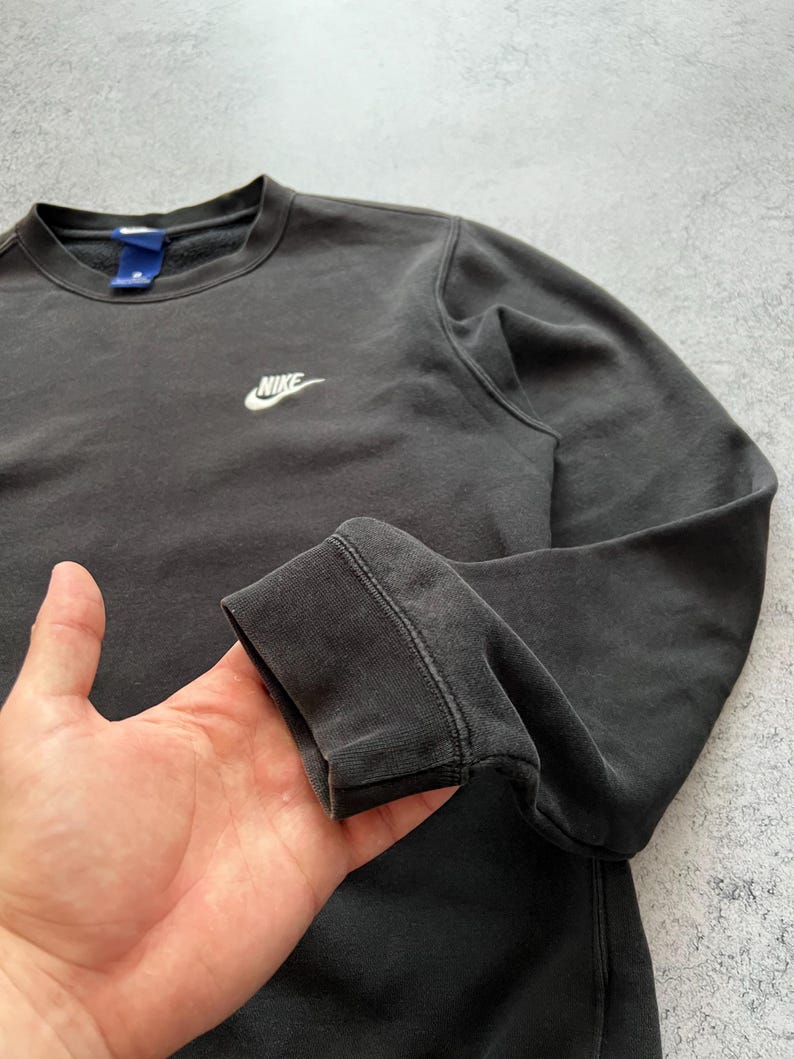 May include: Dark gray crew neck sweatshirt with the Nike logo embroidered in white on the chest. The sweatshirt has long sleeves and a ribbed cuff. The fabric appears to be slightly faded, giving it a vintage look. The collar has a blue tag.