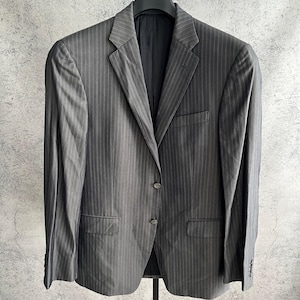 May include: A charcoal gray blazer featuring a subtle vertical pinstripe pattern, a notched lapel, two front buttons, a single chest pocket, and two flap pockets at the waist, displayed on a black hanger against a textured gray background.