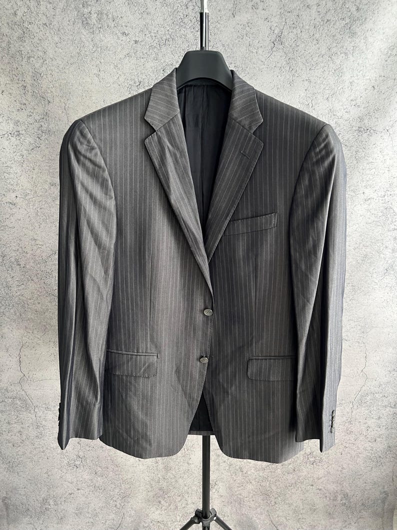 May include: A charcoal gray blazer featuring a subtle vertical pinstripe pattern, a notched lapel, two front buttons, a single chest pocket, and two flap pockets at the waist, displayed on a black hanger against a textured gray background.