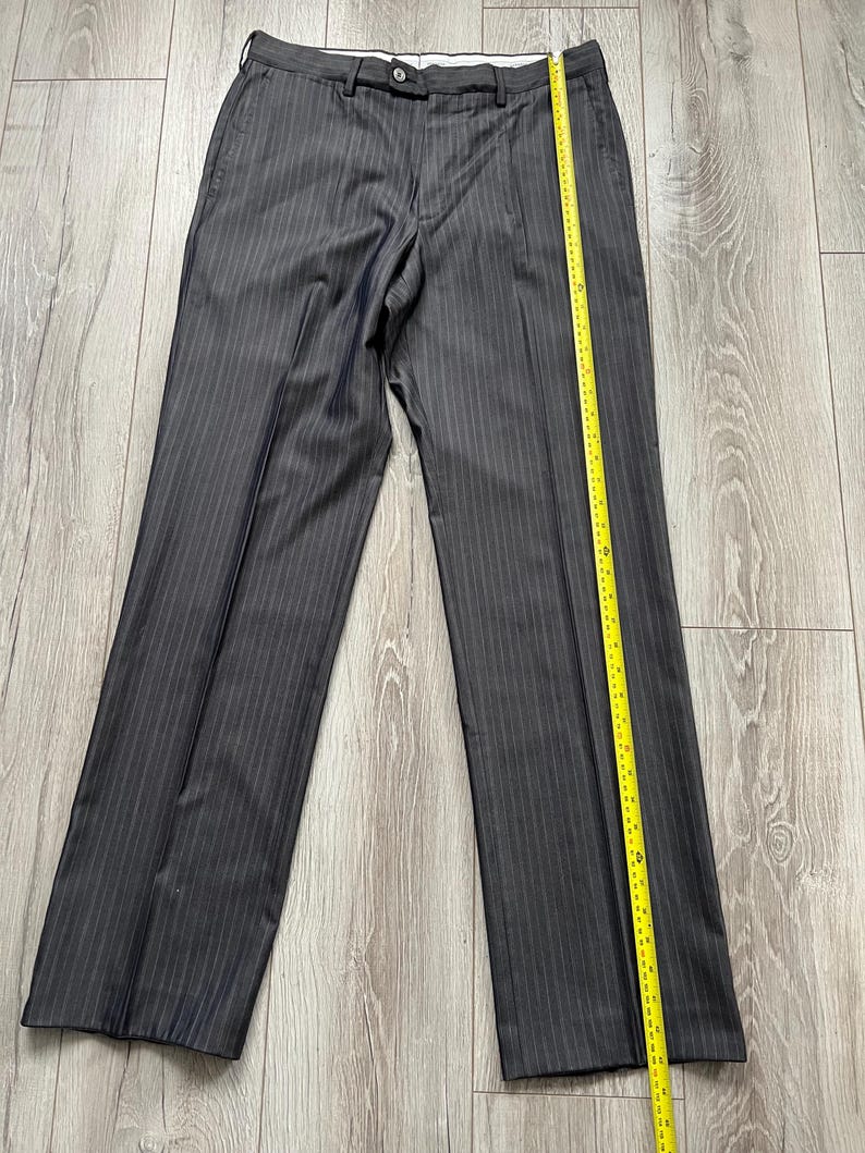 May include: Dark gray trousers with a subtle vertical pinstripe pattern, featuring a button closure at the waistband and a yellow measuring tape placed along the right leg, showing a length of approximately 42 inches from waist to hem.